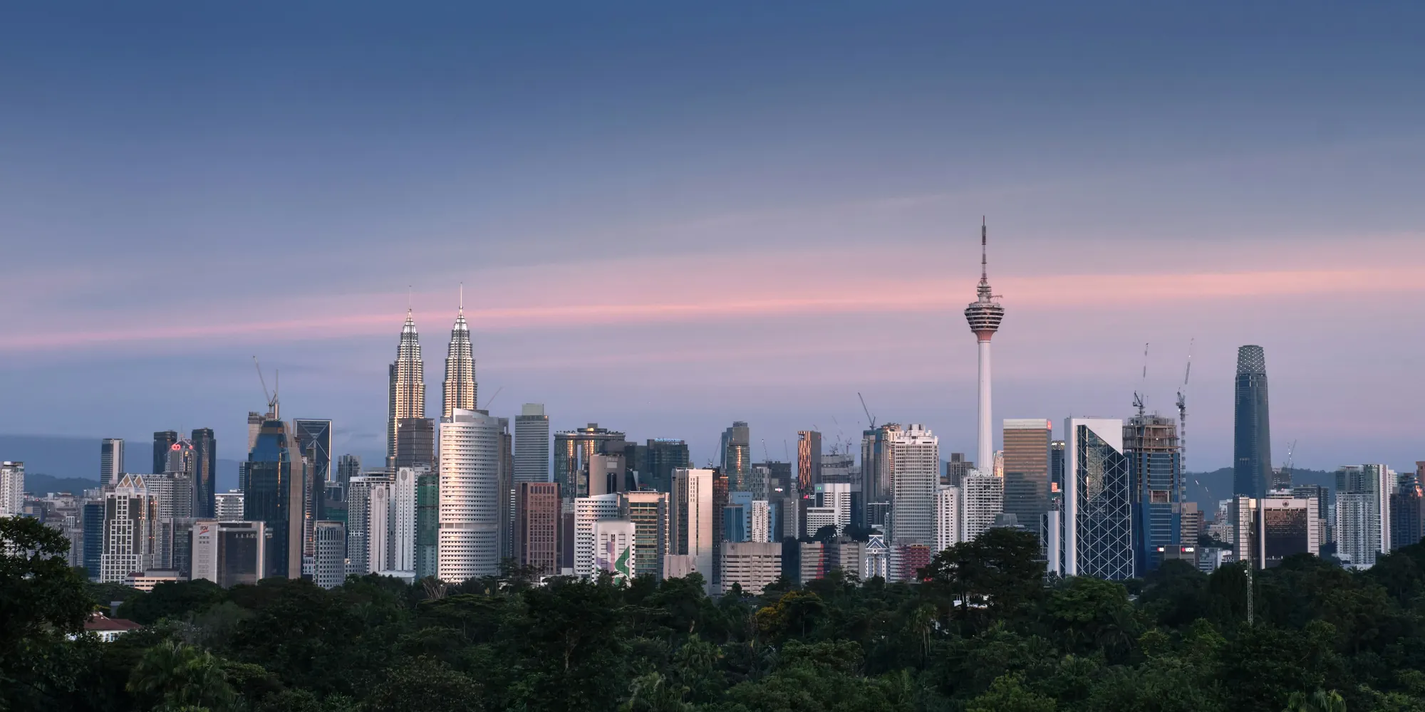 Latar langit Kuala Lumpur waktu senja, dengan Menara Berkembar Petronas dan Menara KL di atas hutan hijau